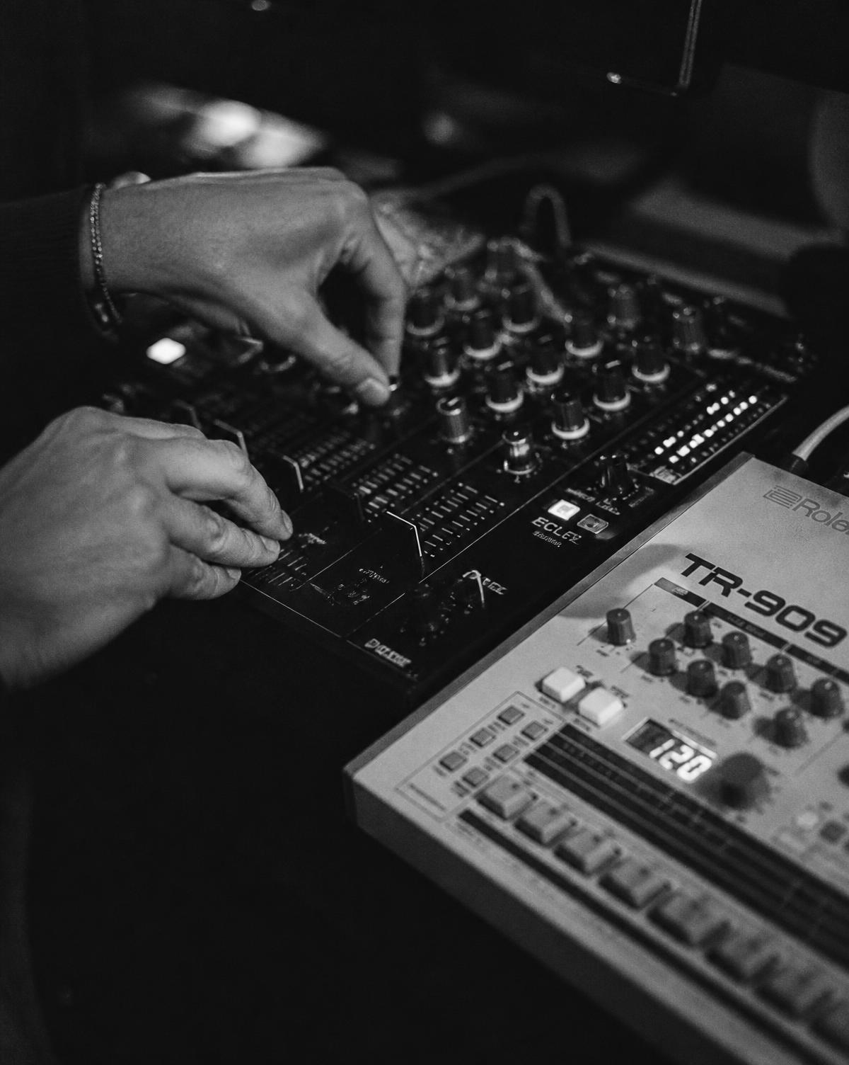Close-up of hands on an analogue DJ mixer beside a TR-909 drum machine